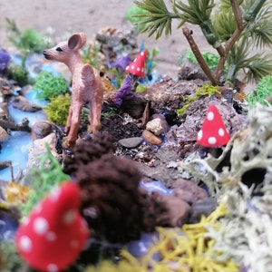 May include: A miniature woodland scene featuring a small deer figurine, red mushrooms with white spots, and a stream. The scene includes miniature trees, moss, and rocks, creating a detailed diorama. The color palette includes shades of brown, green, red, and blue.
