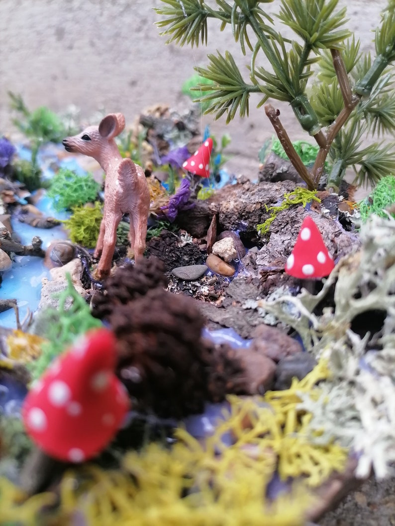 May include: A miniature woodland scene featuring a small deer figurine, red mushrooms with white spots, and a stream. The scene includes miniature trees, moss, and rocks, creating a detailed diorama. The color palette includes shades of brown, green, red, and blue.