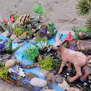 May include: A miniature woodland diorama featuring a stream with blue resin water, rocks, and greenery. A small fawn with white spots stands near the water. Red and white mushroom accents add a whimsical touch to this nature-themed display.