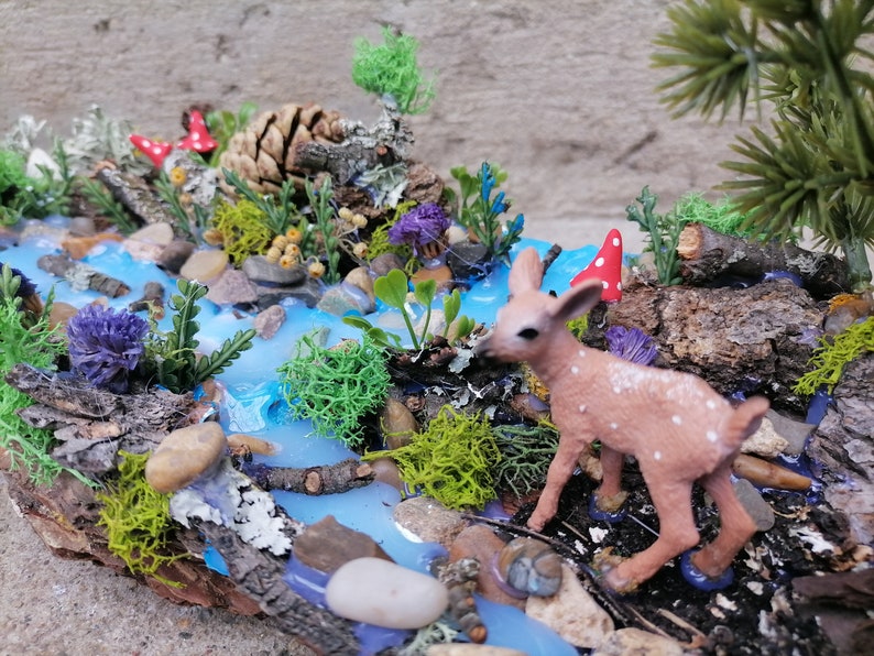 May include: A miniature woodland diorama featuring a stream with blue resin water, rocks, and greenery. A small fawn with white spots stands near the water. Red and white mushroom accents add a whimsical touch to this nature-themed display.