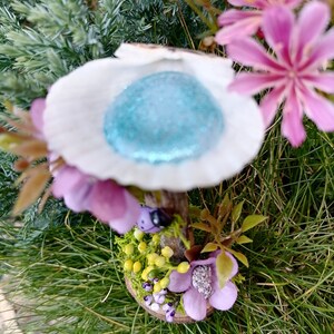 Op de afbeelding: Een miniatuur fee&euml;ntuin met een witte, schelpvormige structuur met een blauwe glittertop, een houten steel en een basis versierd met groen mos, gele bloemen en paarse bloemen.