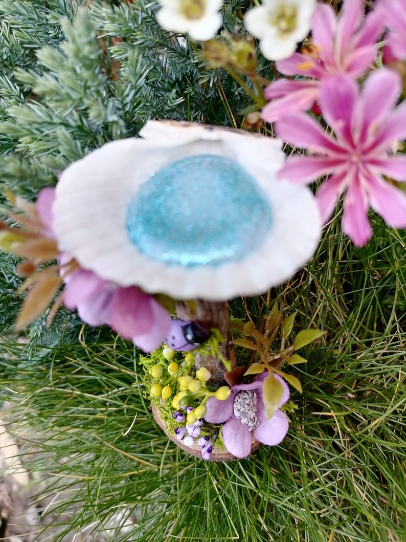 Op de afbeelding: Een miniatuur fee&euml;ntuin met een witte, schelpvormige structuur met een blauwe glittertop, een houten steel en een basis versierd met groen mos, gele bloemen en paarse bloemen.