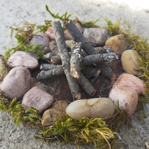 Pu&ograve; includere: Una scena di fal&ograve; in miniatura con piccoli ramoscelli disposti in cerchio su una base marrone, circondata da un anello di pietre rosa chiaro e marroni, il tutto incastonato nel muschio verde.
