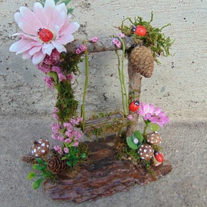 May include: A miniature fairy garden swing set made of twigs and moss with a pink flower, green leaves, and red ladybugs.