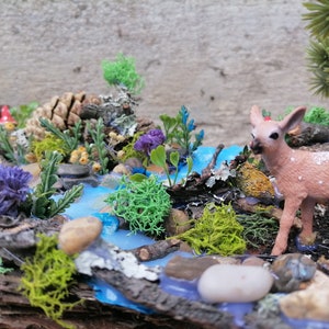 May include: A miniature woodland diorama featuring a small fawn with white spots near a blue stream. The scene includes miniature plants, rocks, and a pinecone, creating a naturalistic, decorative display.