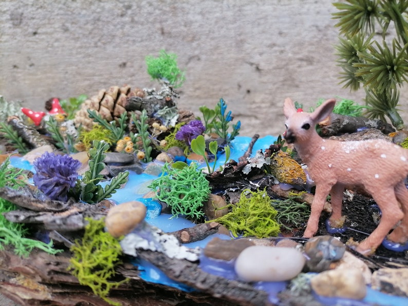 May include: A miniature woodland diorama featuring a small fawn with white spots near a blue stream. The scene includes miniature plants, rocks, and a pinecone, creating a naturalistic, decorative display.