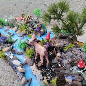 May include: A miniature woodland diorama featuring a small deer figurine near a blue resin river. The scene includes miniature trees, rocks, moss, and red mushroom accents, creating a whimsical forest setting.
