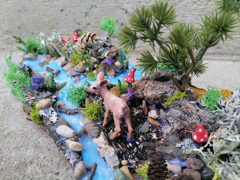May include: A miniature woodland diorama featuring a small deer figurine near a blue resin river. The scene includes miniature trees, rocks, moss, and red mushroom accents, creating a whimsical forest setting.