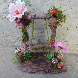 May include: A miniature fairy garden swing set made of twigs and moss, with pink flowers, red ladybugs, and mushrooms.
