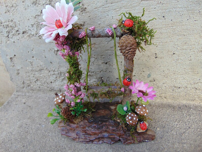May include: A miniature fairy garden swing set made of twigs and moss, with pink flowers, red ladybugs, and mushrooms.