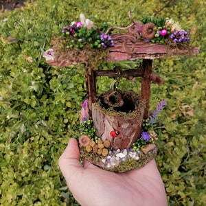Könnte beinhalten: Ein Miniatur-Brunnen aus Holz mit einem moosbewachsenen Dach und einem Eimer, der an einem Seil hängt. Der Brunnen ist mit kleinen Blumen, Eicheln und einem Marienkäfer verziert.