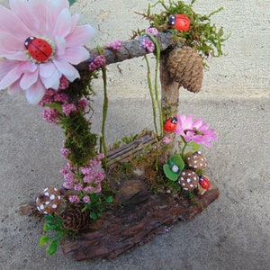 May include: A miniature fairy garden swing set made from twigs and moss, decorated with pink flowers, ladybugs, and mushrooms.