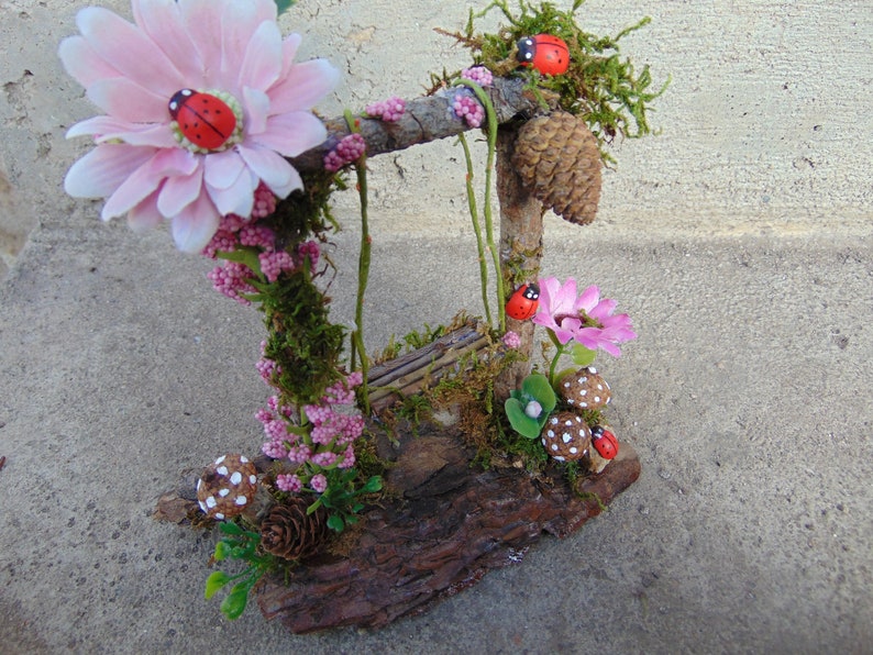 May include: A miniature fairy garden swing set made from twigs and moss, decorated with pink flowers, ladybugs, and mushrooms.