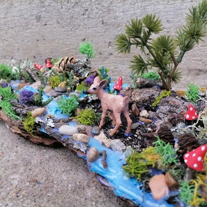 May include: A miniature woodland diorama featuring a small deer figurine near a blue stream. The scene includes miniature trees, red and white spotted mushrooms, and various types of moss and foliage, all arranged on a piece of bark.