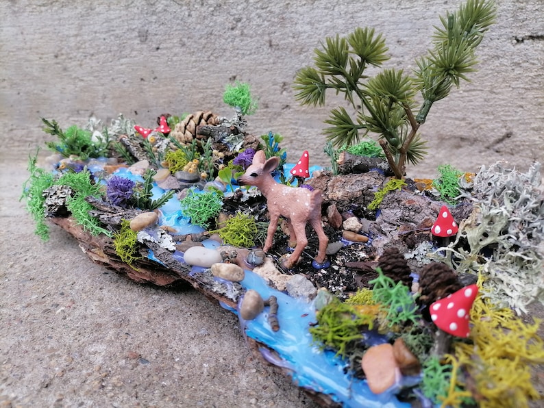 May include: A miniature woodland diorama featuring a small deer figurine near a blue stream. The scene includes miniature trees, red and white spotted mushrooms, and various types of moss and foliage, all arranged on a piece of bark.