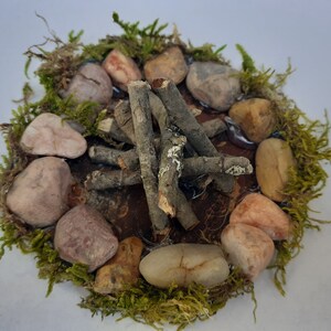 Pu&ograve; includere: Una scena di fal&ograve; in miniatura con bastoncini marroni disposti a forma di teepee, circondati da un anello di pietre di vari colori e muschio verde.