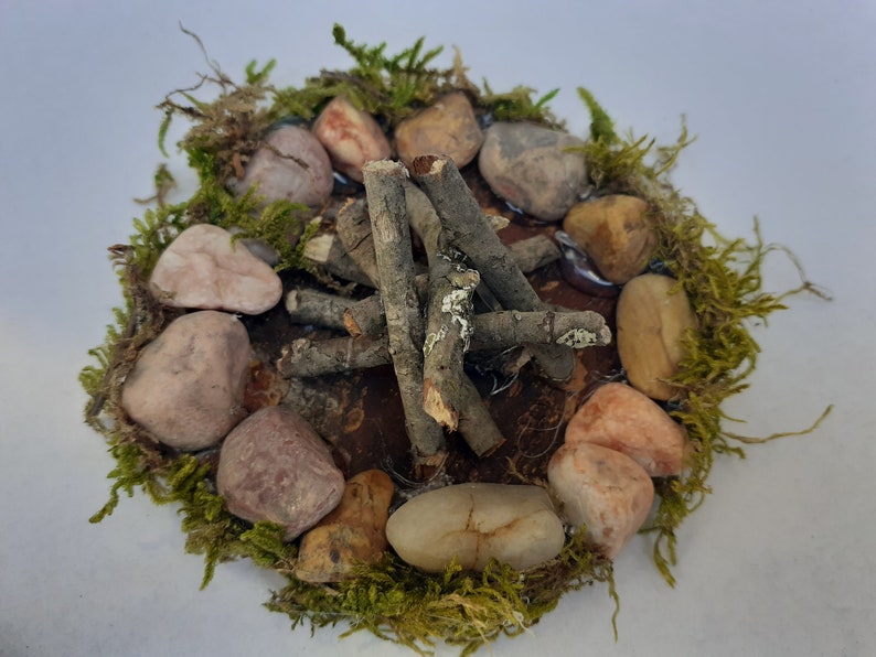 Pu&ograve; includere: Una scena di fal&ograve; in miniatura con bastoncini marroni disposti a forma di teepee, circondati da un anello di pietre di vari colori e muschio verde.