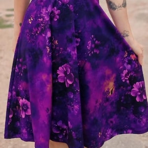 May include: A woman wearing a purple floral print dress with a V-neckline and short sleeves. The dress has a fitted bodice and a flowing skirt.