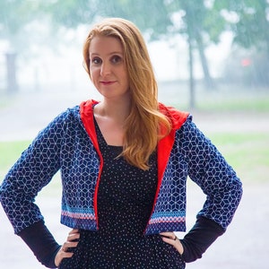 May include: A woman wearing a blue and white geometric patterned cropped jacket with a red hood. The jacket has a colorful trim around the bottom edge. She is wearing a black polka dot dress and has long red hair.