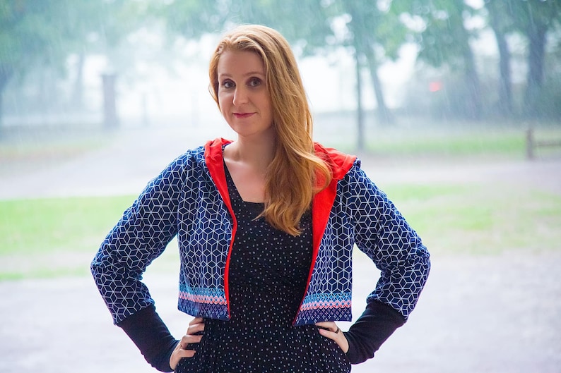 May include: A woman wearing a blue and white geometric patterned cropped jacket with a red hood. The jacket has a colorful trim around the bottom edge. She is wearing a black polka dot dress and has long red hair.