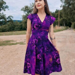 May include: A woman wearing a purple floral print dress with a V-neckline and short sleeves. The dress has a fitted bodice and a flowing skirt.