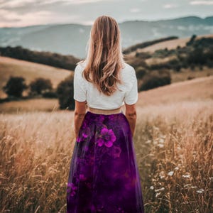 May include: A woman wearing a white shirt and a long purple skirt with a floral print stands in a field of tall grass. The skirt has a dark purple background with pink and white flowers.