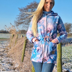May include: A floral print jacket in shades of blue and pink, featuring a hood, pockets, and a drawstring waist. The jacket is worn with blue jeans. The background shows a field and a fence.