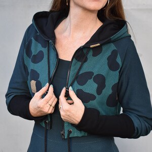 May include: A teal green cropped hoodie with black leopard print and black sleeves. The hoodie has a black zipper and a drawstring.