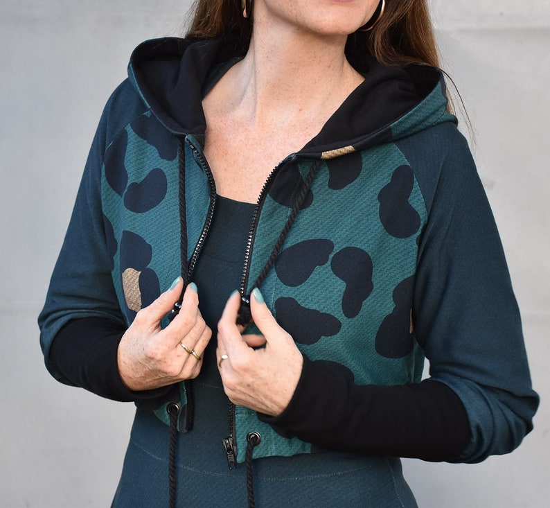 May include: A teal green cropped hoodie with black leopard print and black sleeves. The hoodie has a black zipper and a drawstring.