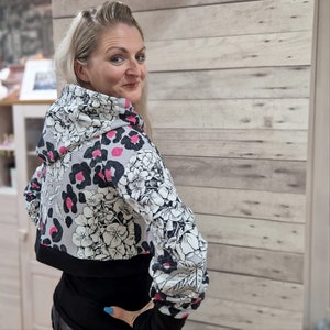 May include: A cropped hoodie with a black and white floral print and pink leopard spots. The hoodie has a black hood and a black band around the bottom.