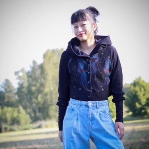 May include: A black cropped hoodie with a blue and red bird design and a row of silver buttons. The hoodie has a hood and long sleeves. The model is wearing blue jeans.
