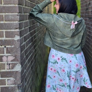 May include: A woman wearing a green hooded jacket with a pink bow on the back and a floral skirt.
