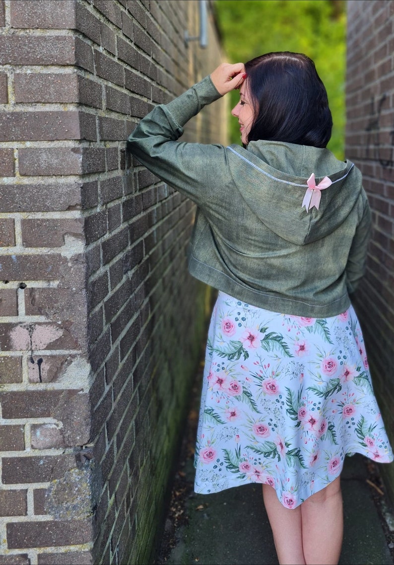 May include: A woman wearing a green hooded jacket with a pink bow on the back and a floral skirt.