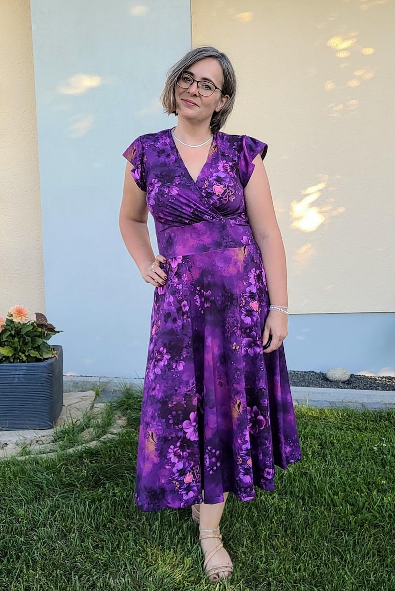 May include: A woman wearing a purple floral print dress with a V-neckline and short sleeves. She is standing in front of a light blue and yellow wall.