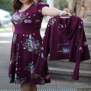 May include: A woman is holding a maroon hooded sweatshirt with a floral print and a sewing machine graphic. The sweatshirt has long sleeves and a relaxed fit. She is also wearing a maroon floral dress with a fitted bodice and a full skirt.