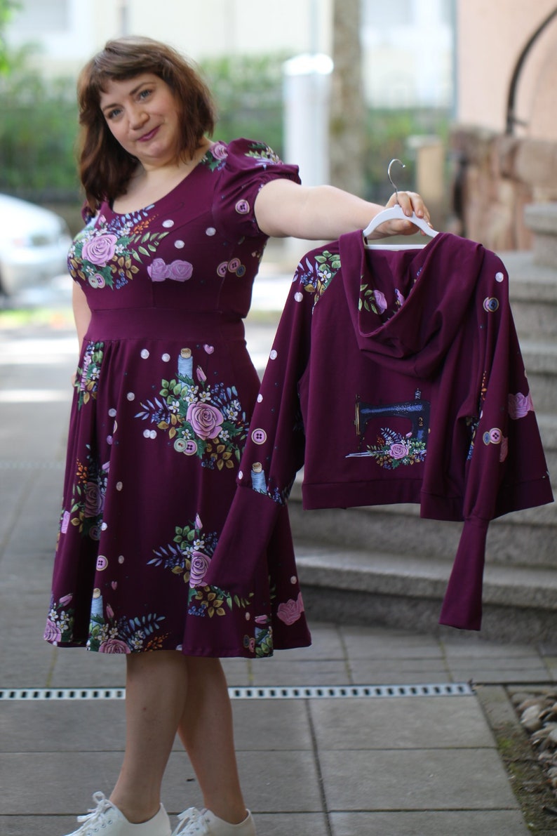 May include: A woman is holding a maroon hooded sweatshirt with a floral print and a sewing machine graphic. The sweatshirt has long sleeves and a relaxed fit. She is also wearing a maroon floral dress with a fitted bodice and a full skirt.