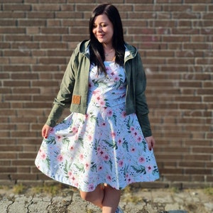 May include: A woman wearing a green hooded jacket over a white dress with pink floral print. The dress has a full skirt and the jacket has a patch on the left sleeve that says "LOVE".