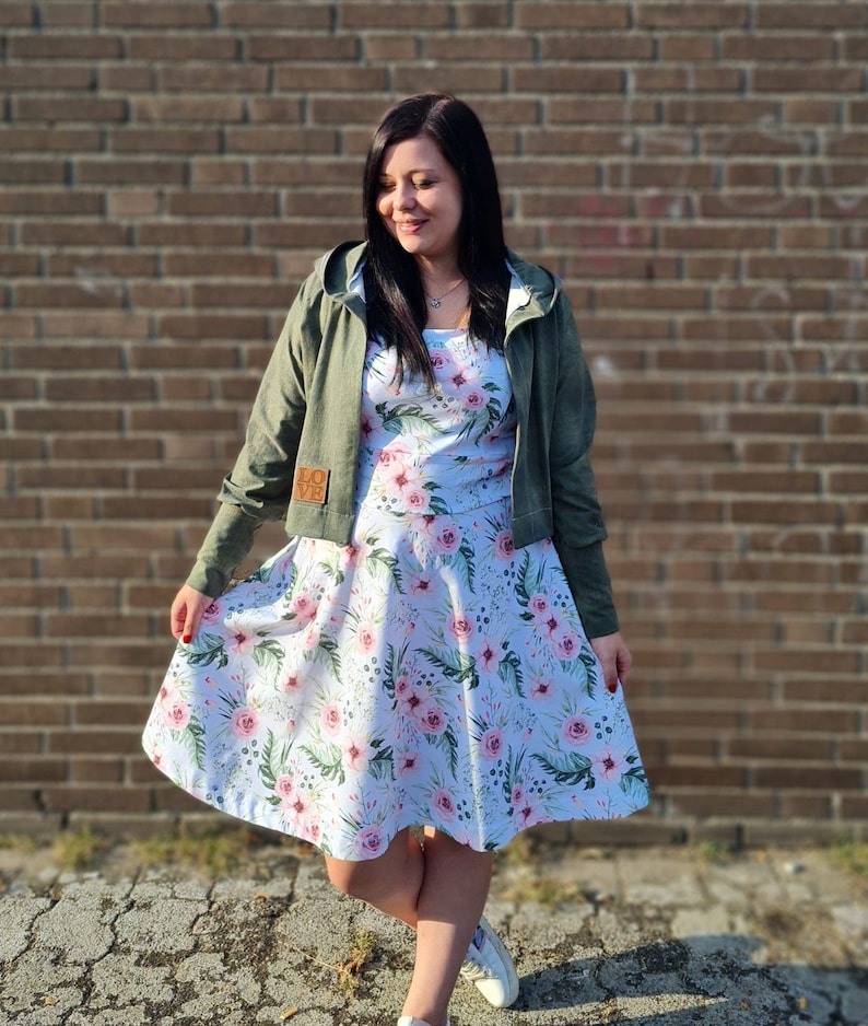 May include: A woman wearing a green hooded jacket over a white dress with pink floral print. The dress has a full skirt and the jacket has a patch on the left sleeve that says "LOVE".