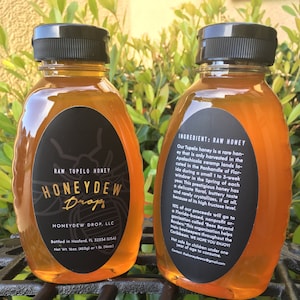May include: Two glass bottles of raw Tupelo honey, labeled "Honeydew Drop" with a black and gold label. The bottles are sitting on a black metal grate. The label on the right bottle reads "Ingredient: Raw Honey" and includes information about the honey's origin and the nonprofit organization that benefits from its sales.