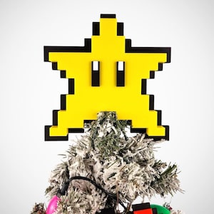 May include: A yellow pixelated star with black outlines and a face, used as a Christmas tree topper. The star is on top of a small artificial Christmas tree with white flocked branches and colorful lights.