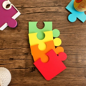 May include: Four colorful puzzle pieces stacked on top of each other. The pieces are green, yellow, orange, and red. The pieces are on a wooden surface.