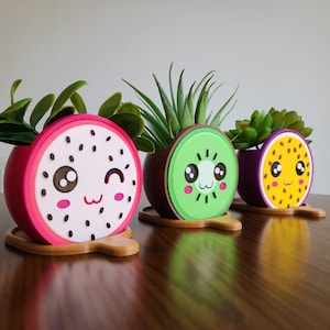 Puede incluir: Tres lindas macetas con forma de fruta con caras de dibujos animados. Las macetas son de color rosa, verde y morado y presentan un diseño de fruta de dragón, kiwi y maracuyá. Cada maceta tiene una pequeña planta creciendo dentro.