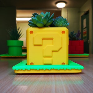 May include: A yellow question mark block planter with a green rim and a succulent plant growing inside. The planter is sitting on a wooden surface.