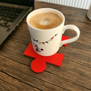 May include: A white ceramic mug with a black and red design of flags and people on a string. The mug is filled with coffee and is sitting on a red puzzle-shaped coaster.