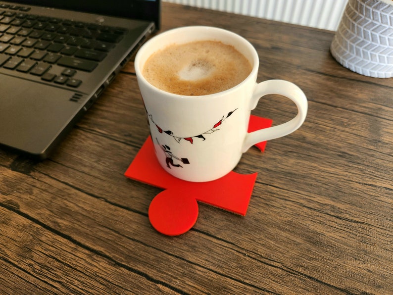 May include: A white ceramic mug with a black and red design of flags and people on a string. The mug is filled with coffee and is sitting on a red puzzle-shaped coaster.