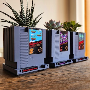 Op de afbeelding: Drie grijze en zwarte Nintendo Entertainment System (NES) gamecartridge-vormige potten met verschillende speltitels. De potten zijn gevuld met vetplanten. De titels zijn "Super Piranha Plant", "Succublaster" en "Zombie".
