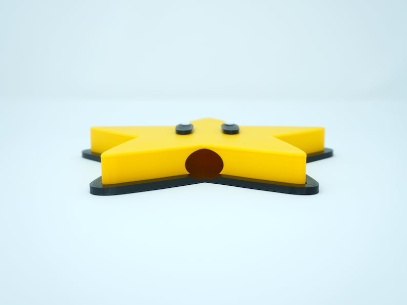 May include: A yellow star-shaped object with two black eyes and a large black mouth. The star is made of plastic and has a black outline.
