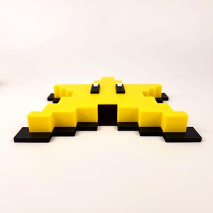 May include: A yellow pixelated star-shaped ornament with black and white details. The ornament is made of plastic and has a flat back.