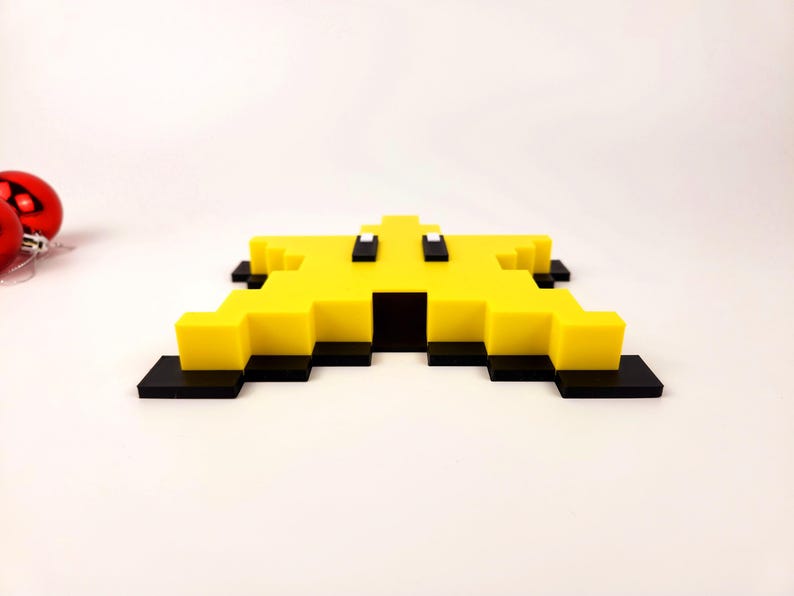 May include: A yellow pixelated star-shaped ornament with black and white details. The ornament is made of plastic and has a flat back.