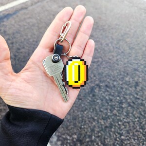May include: A silver key with a black and silver keychain attached to a gold pixelated coin charm.